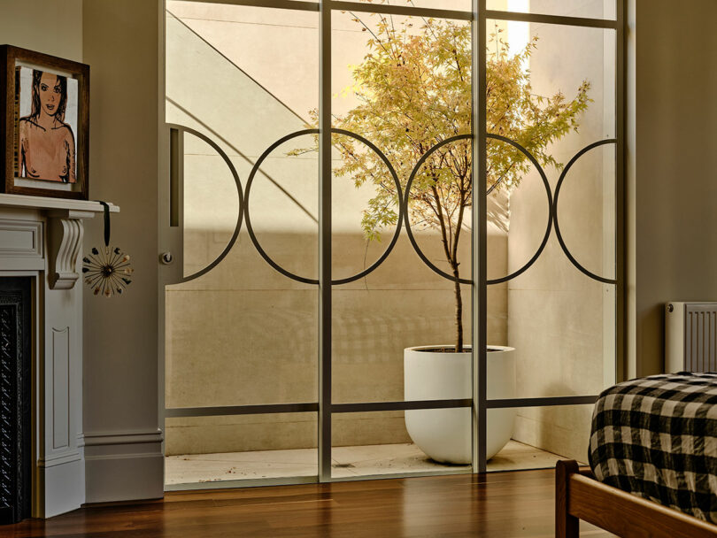 A modern bedroom with a checkered bedspread, a potted tree behind a decorative glass partition, a white fireplace, and a framed portrait on the mantel.