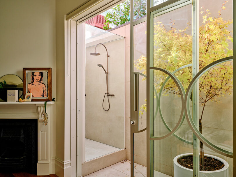 A glass sliding door opens to an outdoor shower area with a potted tree; inside, a fireplace mantel holds framed art and decor items.
