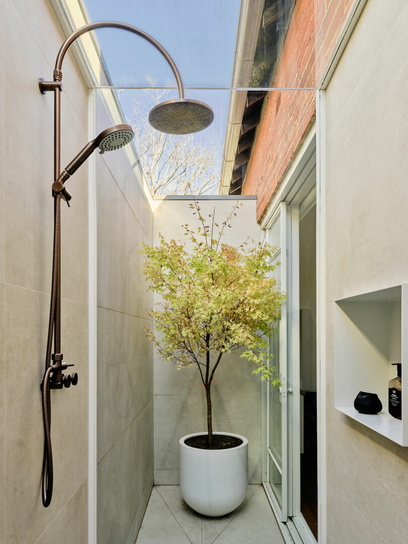 Outdoor shower with a rainfall showerhead, beige tiled walls, a potted tree, and a narrow skylight above letting in natural light.
