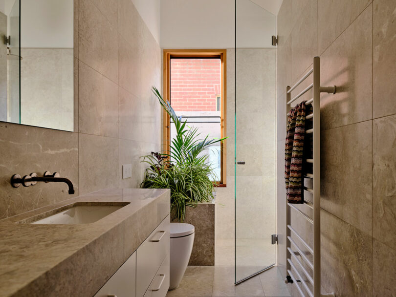 Modern bathroom with beige stone tiles, a glass shower, a white toilet, a wall-mounted sink, a heated towel rack with striped towels, and a potted plant near a window.