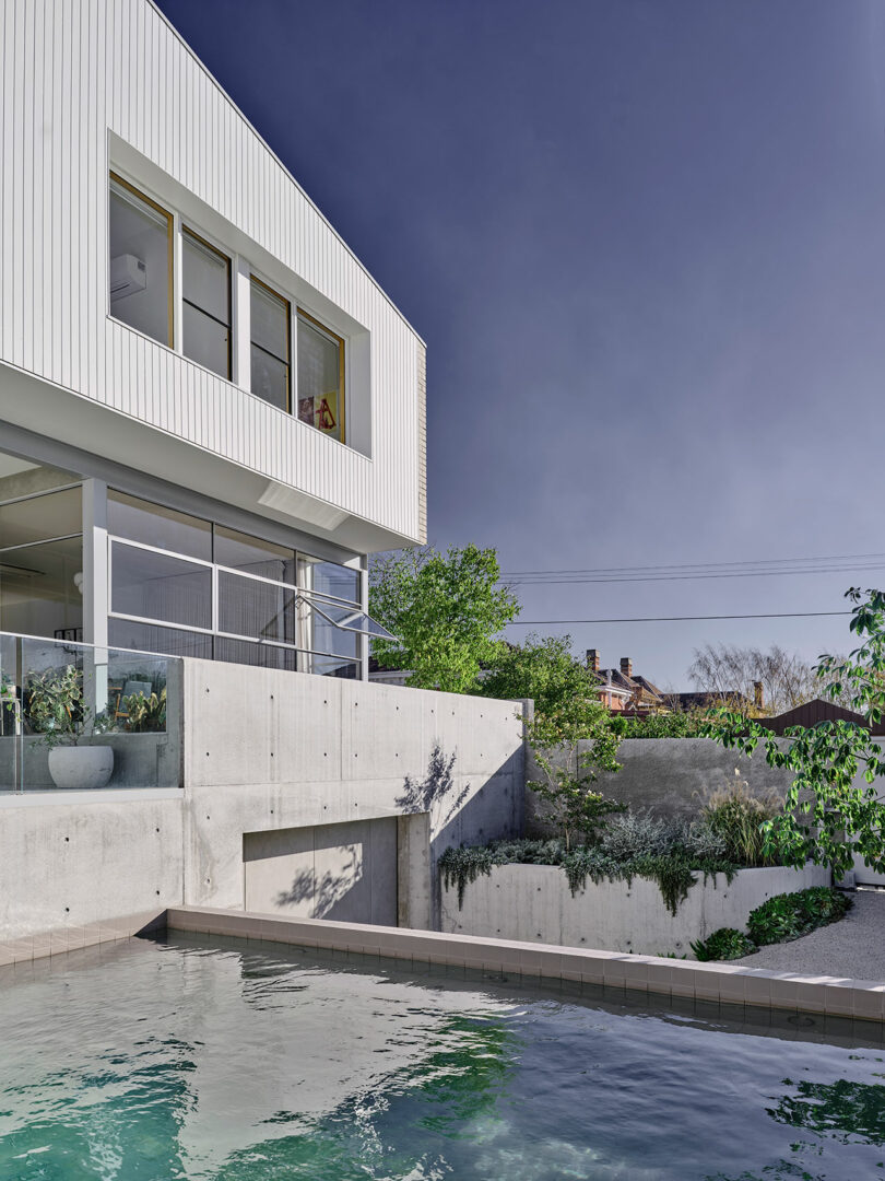 Modern white house with large windows, concrete walls, and a pool in the foreground, surrounded by greenery under a clear sky.