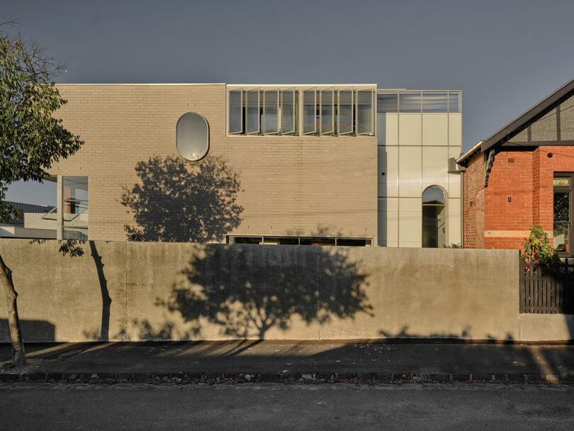 A modern beige building with rectangular windows and a circular window sits behind a concrete wall, casting a tree shadow; a red brick house is partially visible to the right.