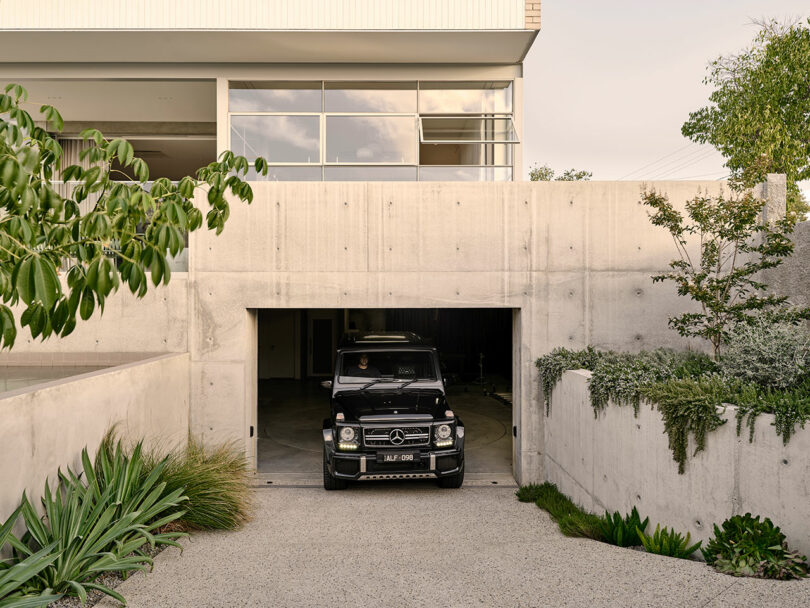 A black Mercedes-Benz G-Class SUV drives out of a modern concrete residential garage surrounded by greenery and plants.