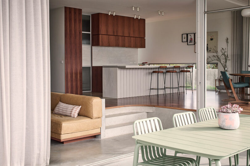 Modern open-plan kitchen and dining area with wood cabinetry, island seating, green outdoor table and chairs, and a tan sofa with a striped pillow.