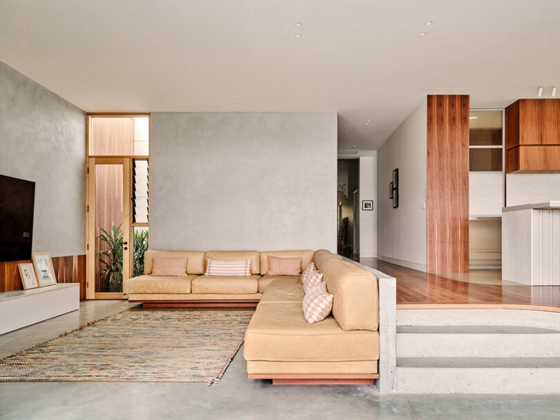A modern living room with a beige sectional sofa, patterned rug, light gray walls, and wooden accents, adjacent to an open kitchen with wood cabinetry.