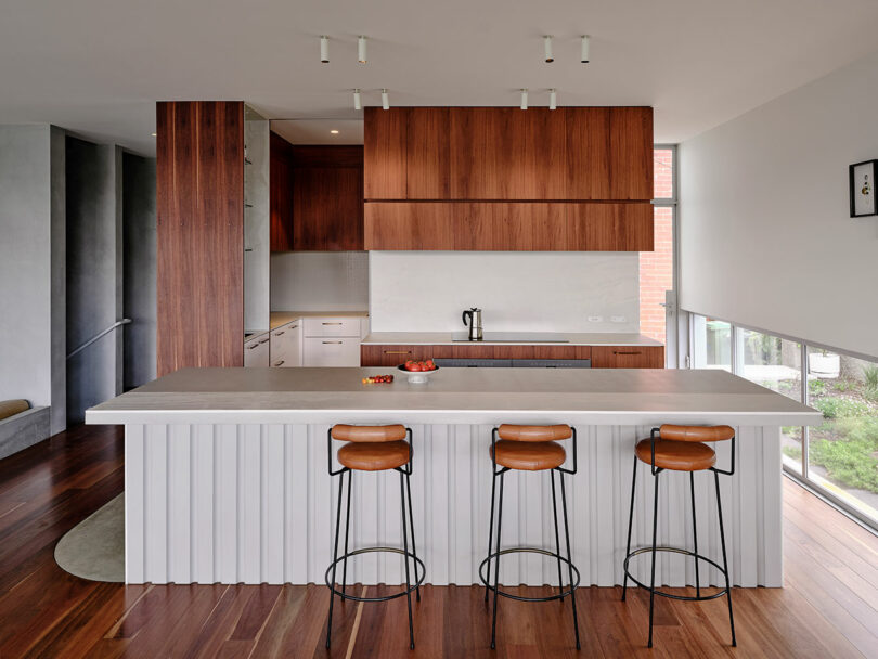 Modern kitchen with wood cabinets, a large island with a ribbed white base, three brown barstools, and floor-to-ceiling windows providing natural light.