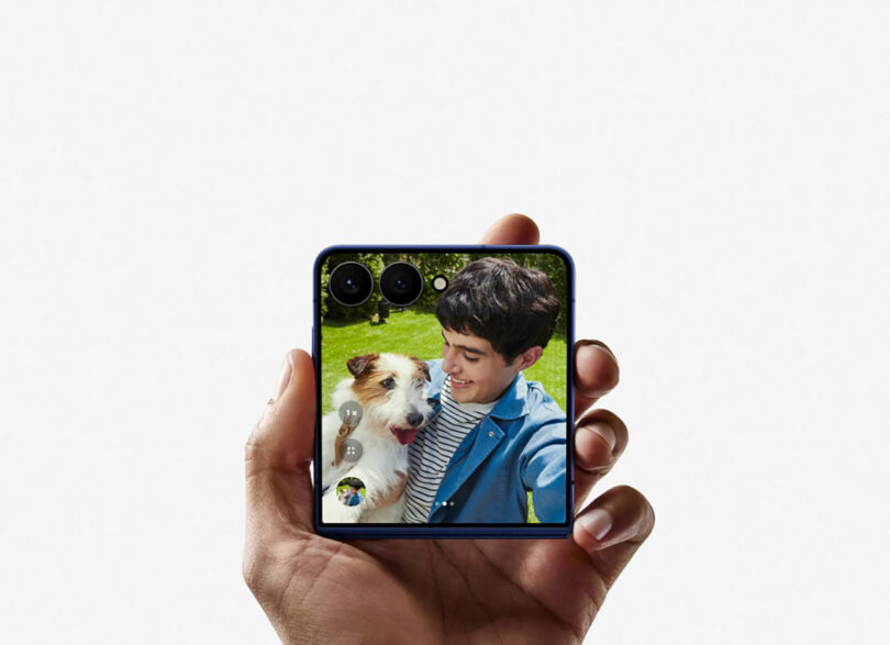 A hand holds a folded Samsung smartphone displaying a photo of a person smiling beside a dog, with trees and grass visible in the background.