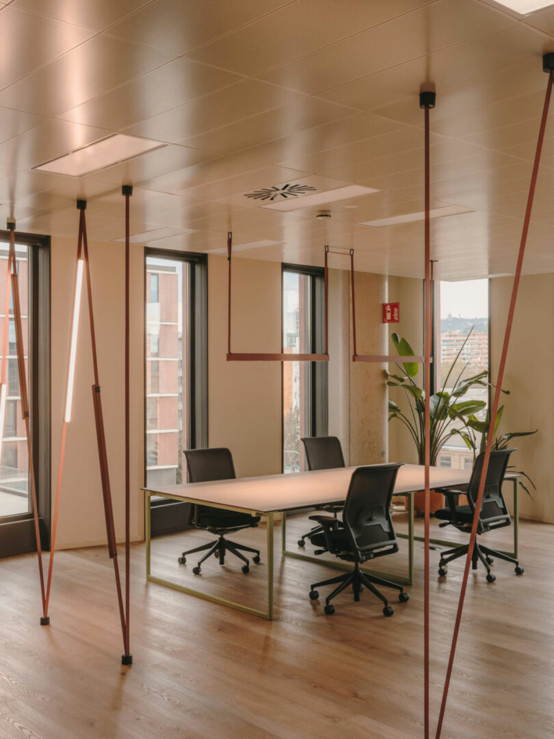 Modern office space with large windows, wooden floor, a rectangular table, four black chairs, overhead lighting, and a potted plant in the corner.