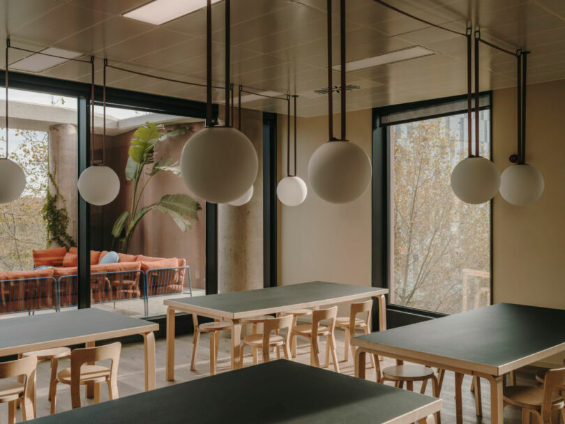 Modern dining area with wooden tables and chairs, large windows, spherical pendant lights, and a view of an outdoor patio with orange seating and greenery.