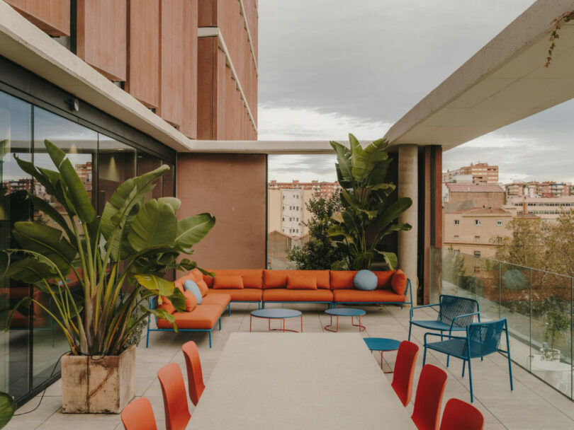Modern rooftop terrace with a long white table, red chairs, orange cushioned seating, blue chairs, potted plants, and a city view in the background.