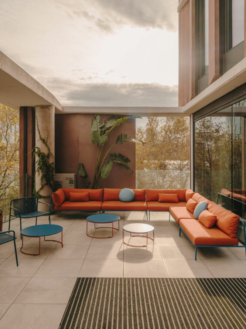 Modern outdoor patio with an L-shaped orange sofa, blue chairs, round tables, large potted plant, glass walls, and neutral tile flooring under a partly cloudy sky.