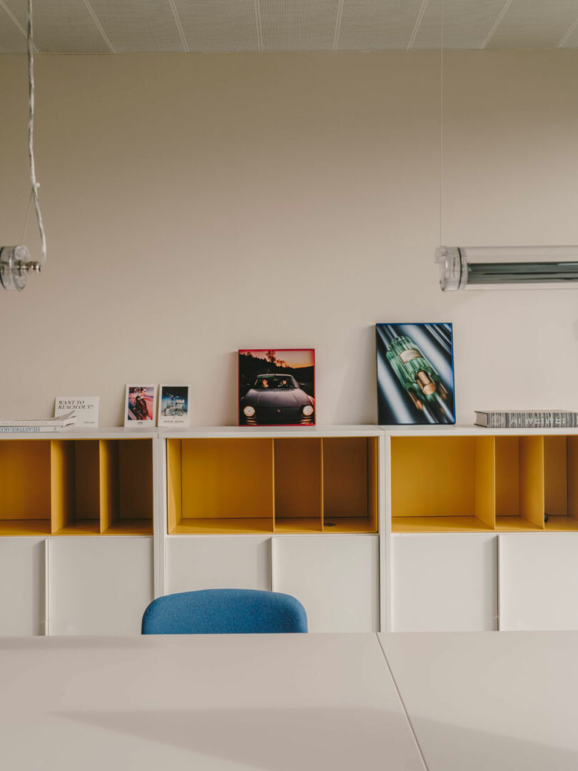 A minimalist office space with a white table, blue chair, yellow-shelved storage units, and framed photos and books arranged on top.