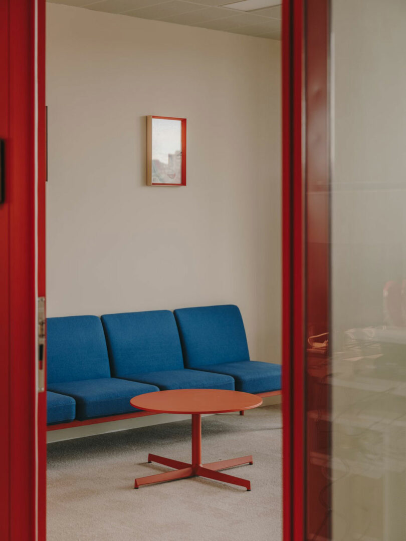 A small office room with a blue sofa, a round red coffee table, and a framed picture on a beige wall, viewed through a doorway with a red frame.