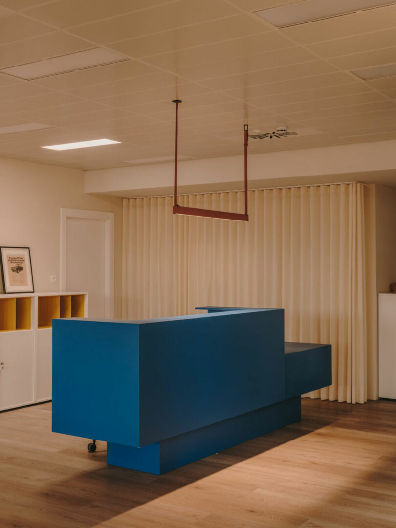 A minimalist office space with a blue geometric reception desk, beige curtains, wooden flooring, and a red ceiling fixture.