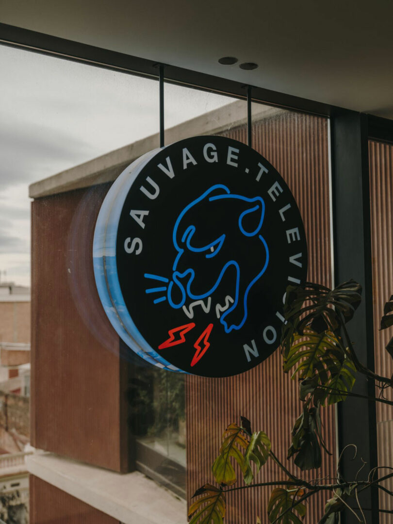A round neon sign with a blue outline of a growling panther, red lightning bolts, and the words "SAUVAGE.TELEVISION" hangs near a window.