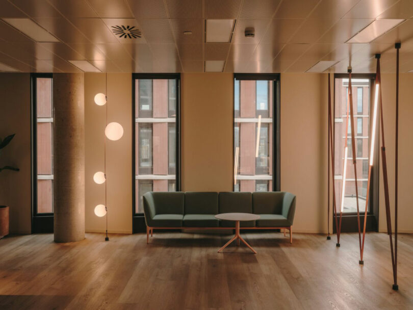 Modern office lounge with a green sofa, round white table, vertical pole lights, and large windows letting in natural light.