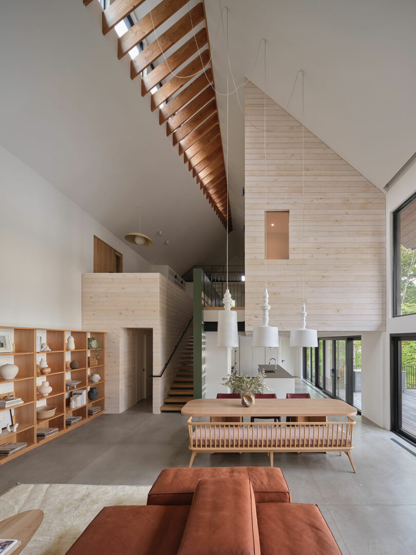 Modern open-concept living space with high ceilings, large windows, wooden accents, and minimalist furniture. Stairs lead to an upper loft area.
