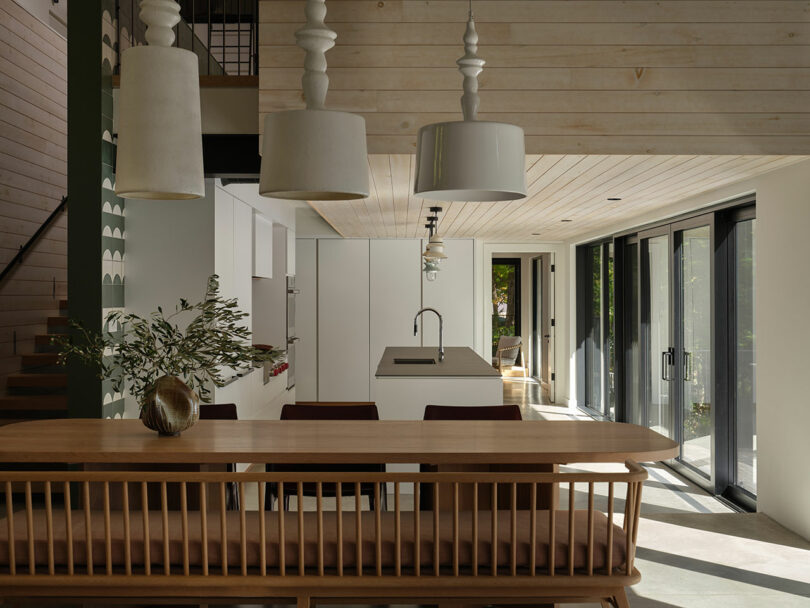 Modern kitchen and dining area with wooden ceiling, large windows, white cabinets, pendant lights, and a wooden table with bench seating. Natural light fills the space.