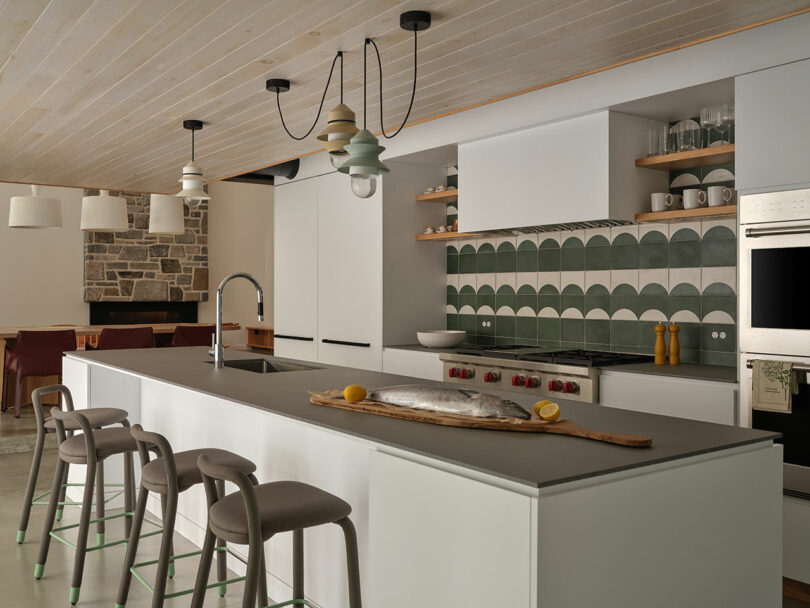 Modern kitchen with a large island, three stools, a whole fish and lemons on a cutting board, green patterned tile backsplash, open shelves, and built-in ovens.