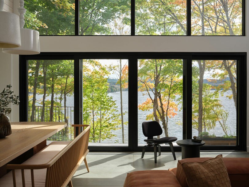 Large windows in a modern living room overlook a lake and trees with autumn foliage; sunlight streams into the minimal, furnished interior.