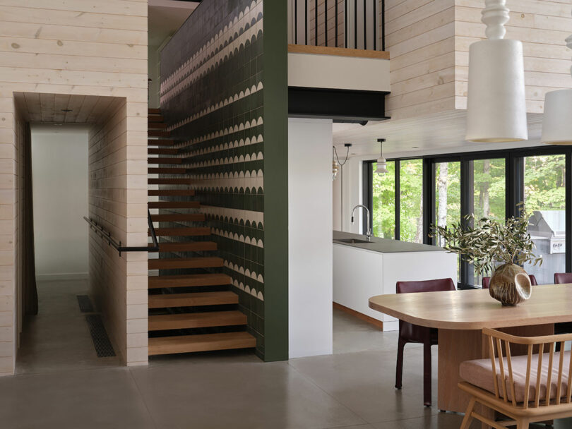 Modern open-concept interior with floating wood staircase, patterned green stair wall, dining area with wooden table and chairs, and a kitchen with large windows.