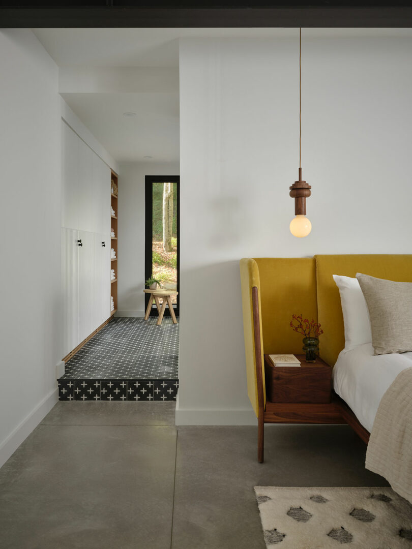 A modern bedroom with a yellow upholstered bed, wooden nightstand, hanging light bulb, and a view into a hallway with patterned tile floor and white walls.