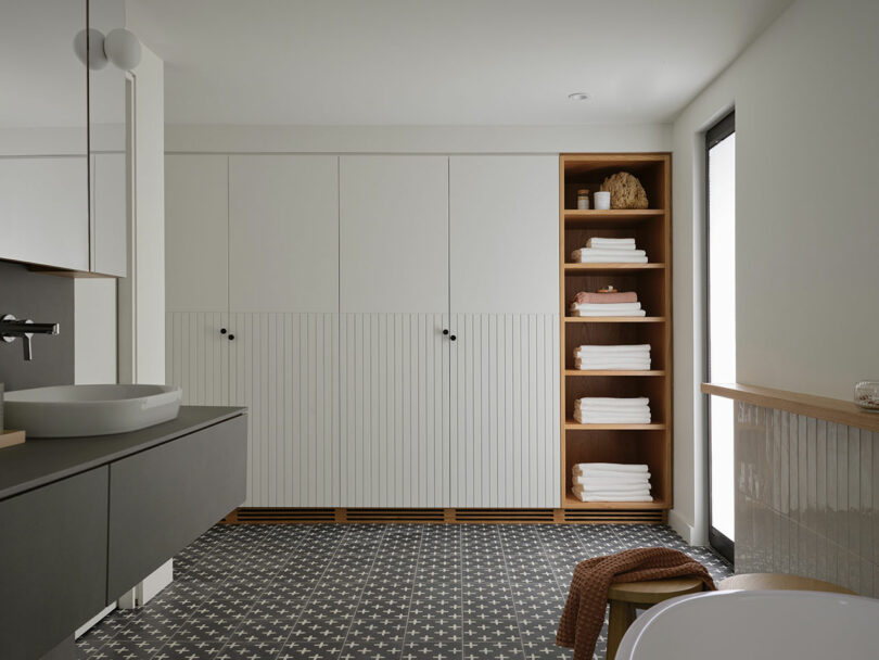 Modern bathroom with black and white patterned floor, minimalist cabinets, open wooden shelving with neatly folded towels, and a round vessel sink on a dark vanity.