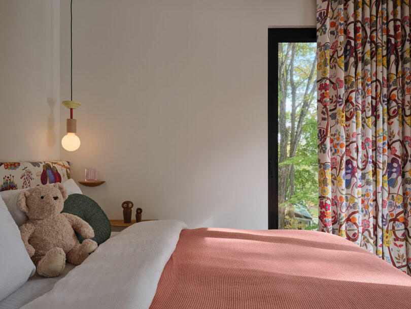 A neatly made bed with a pink blanket, a teddy bear, and cushions is next to a window with floral curtains, overlooking green trees outside. A round light hangs above a small bedside shelf.
