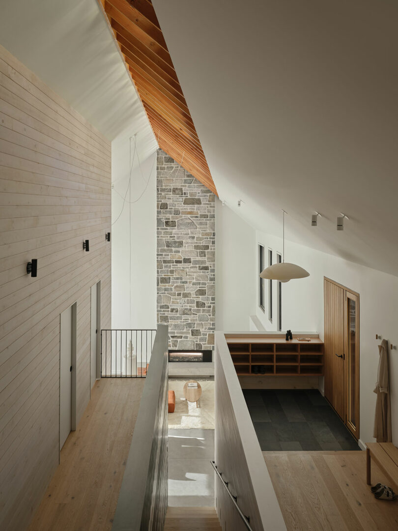 Modern interior with light wood floors, white walls, a stone accent wall, and a sloped ceiling with exposed wooden beams; staircase leads to an entry area with a wooden door.