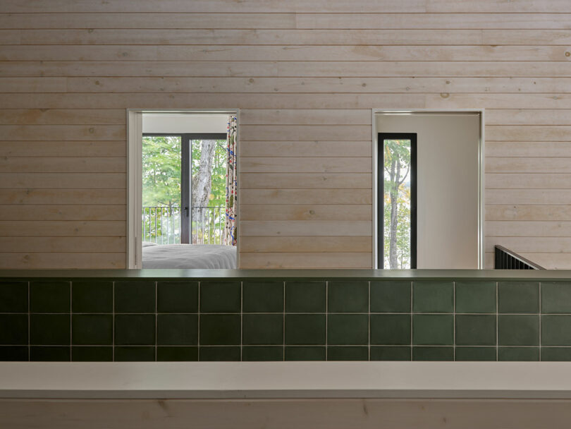 Light wood-paneled interior wall with two open doorways, green tiled half-wall in foreground, and view of trees through windows in the background.