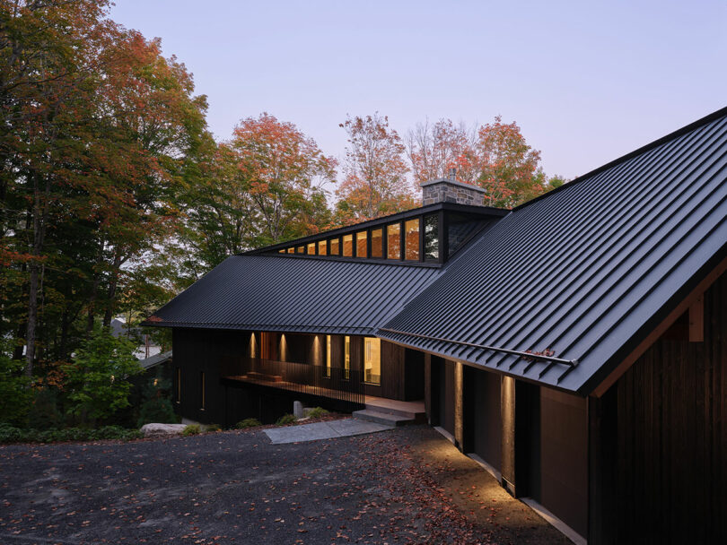 A modern house with dark metal roofing and wood siding, surrounded by trees with autumn foliage, photographed at dusk with interior lights on.