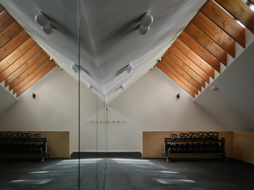 Minimalist gym room with slanted wooden ceiling, mirrored wall, and two racks of kettlebells along opposite walls. Natural light enters from angled windows.