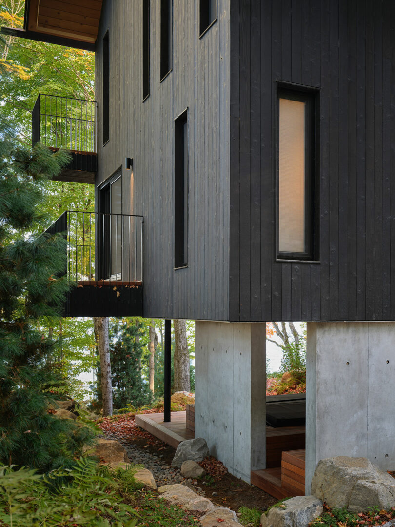 Modern house with black vertical siding and concrete stilts, featuring small balconies and large windows, surrounded by trees and rocks.