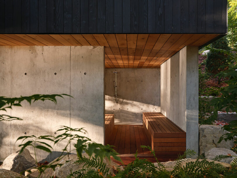 A modern outdoor space with concrete walls, wooden ceiling and bench, and a shower fixture, surrounded by greenery and rocks.