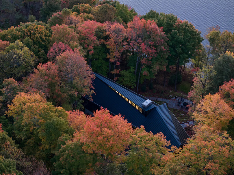 A black-roofed house is nestled among colorful autumn trees near the edge of a lake, viewed from above.