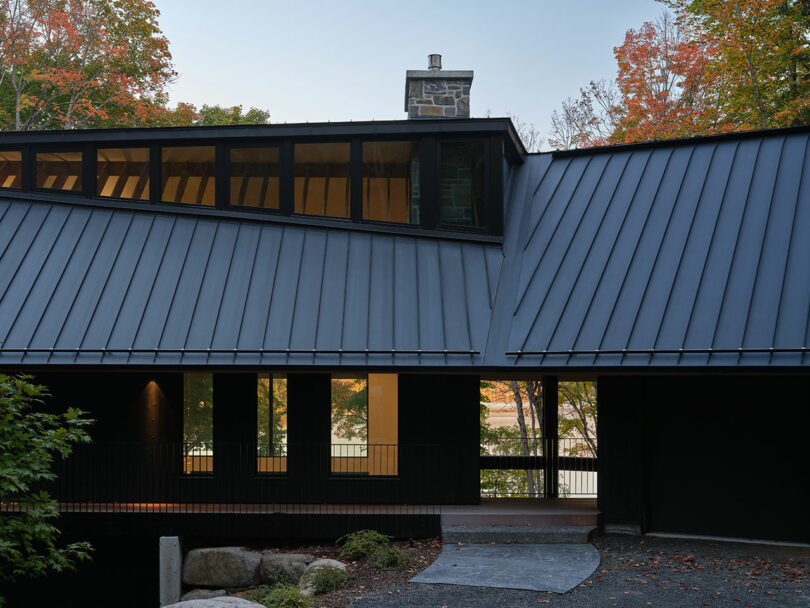A modern building with a dark metal sloped roof, tall windows, and stone chimney, surrounded by trees with autumn foliage.