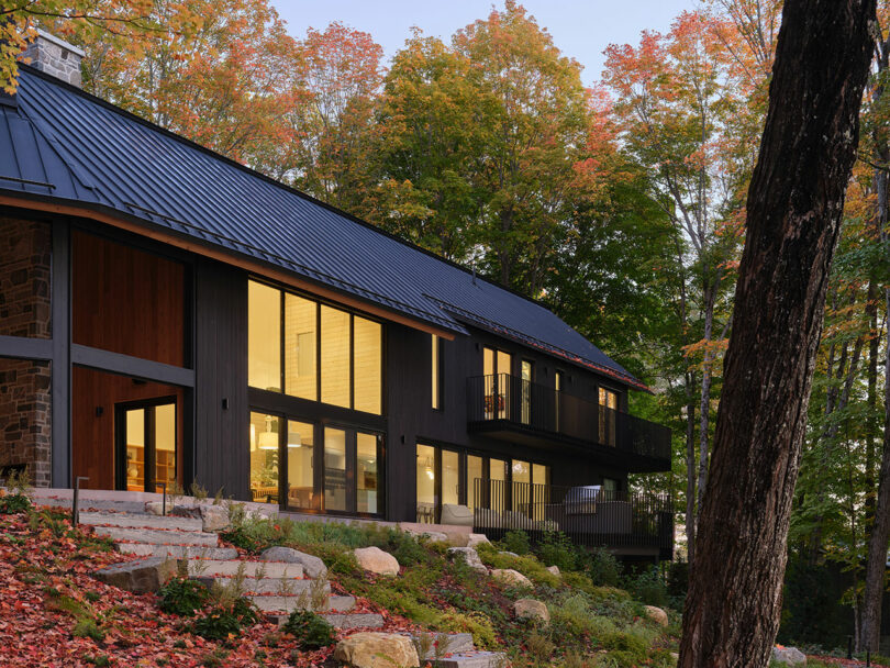 Modern two-story house with large windows and dark wood exterior, surrounded by autumn trees and a rocky, leaf-covered hillside.