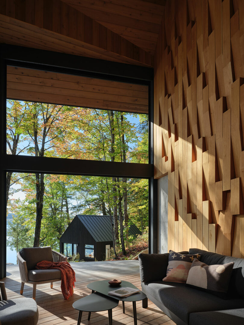 A modern living room with geometric wooden wall panels, large windows overlooking trees, and a small black cabin outside on a deck.