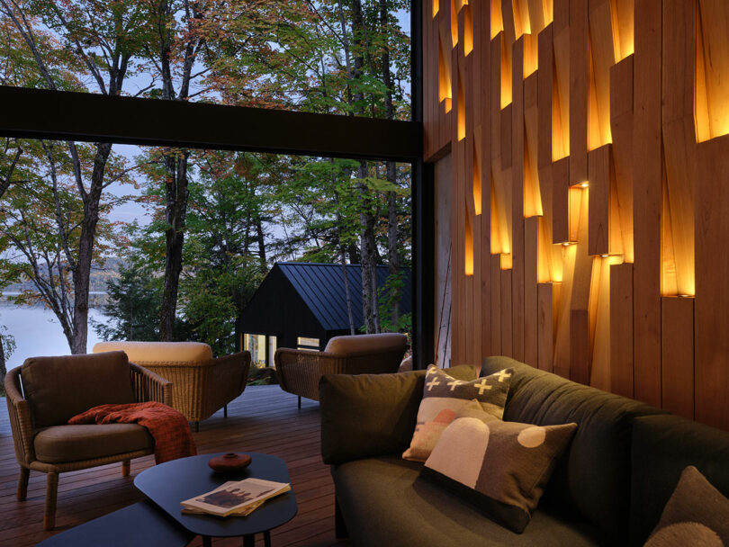 Modern living space with geometric wooden wall lights, contemporary furniture, and a view of a terrace overlooking trees and a lake at dusk.