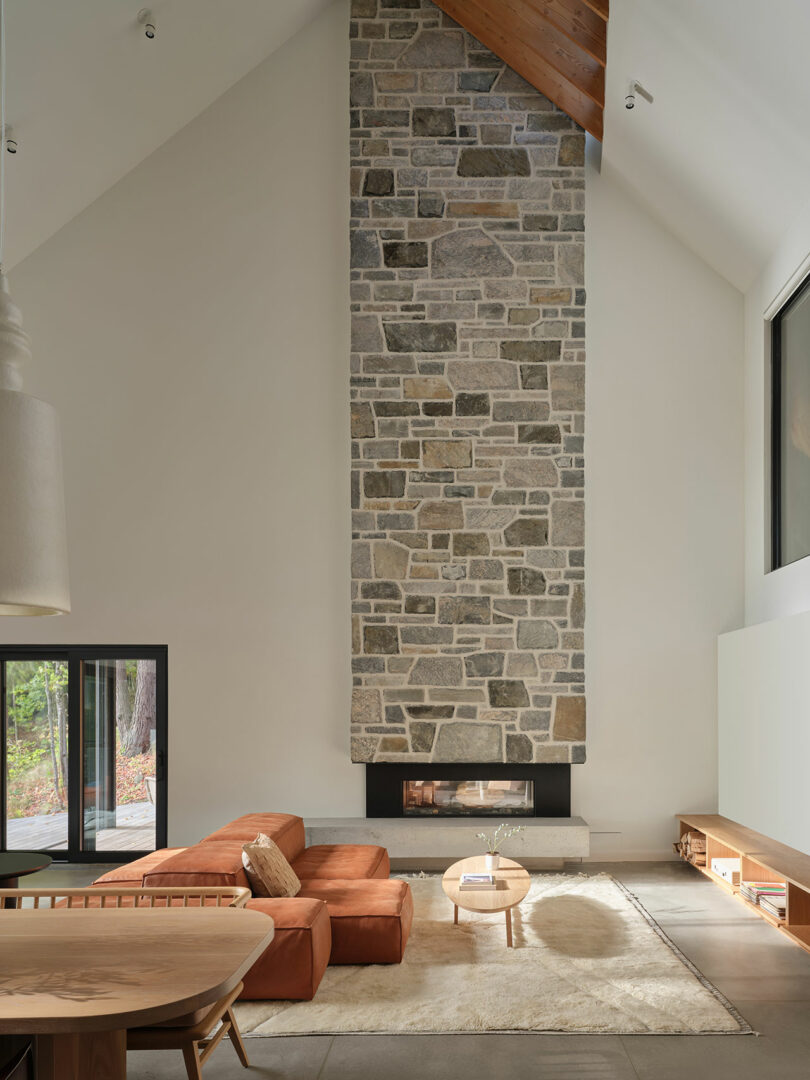 A modern living room with a tall stone fireplace, orange sectional sofa, round wooden coffee table, and large windows letting in natural light.