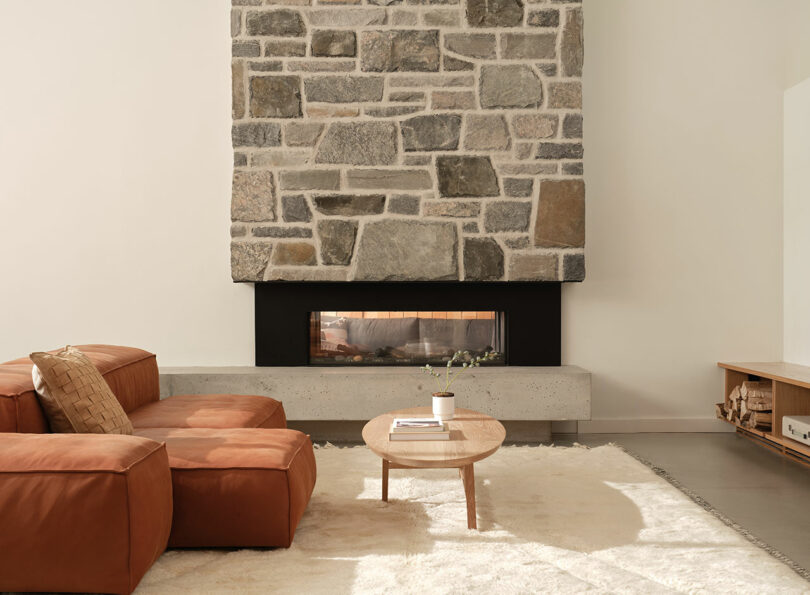 A modern living room with a stone fireplace, brown leather sectional, round wooden coffee table, and white rug on a concrete floor.
