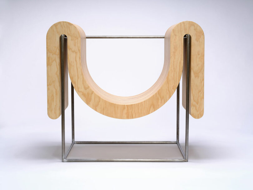 A modern sculpture featuring a large, curved wooden form suspended within a rectangular metal frame, set against a plain white background.