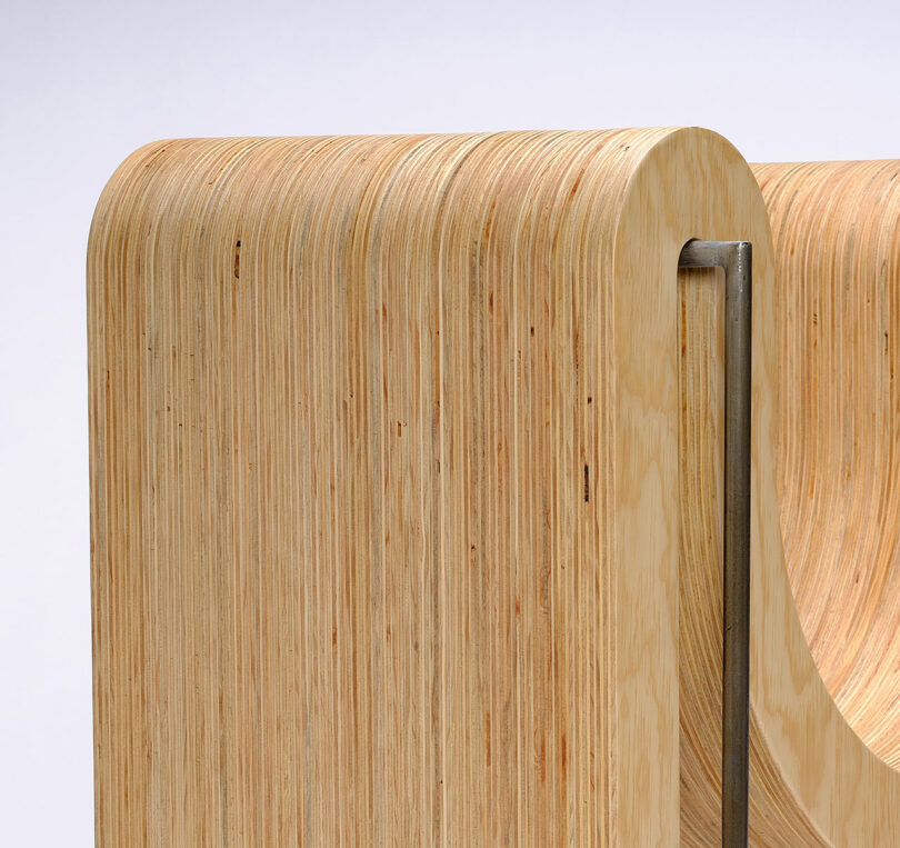 Close-up of a modern wooden structure with smooth, curved layered plywood and a vertical metal support on a plain background.