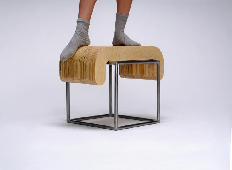 A person wearing gray socks stands on a modern stool made of bent wood and a metal frame, against a plain white background.