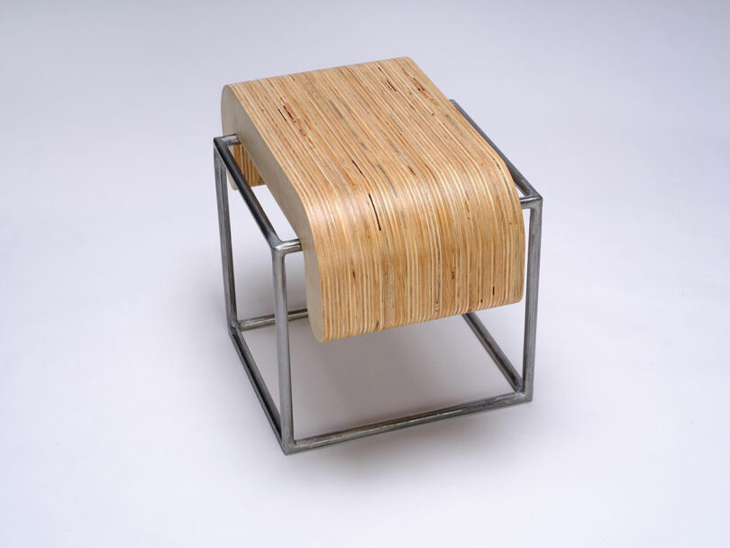 A modern stool with a curved, layered plywood seat resting on a minimalist metal cube frame, set against a plain white background.