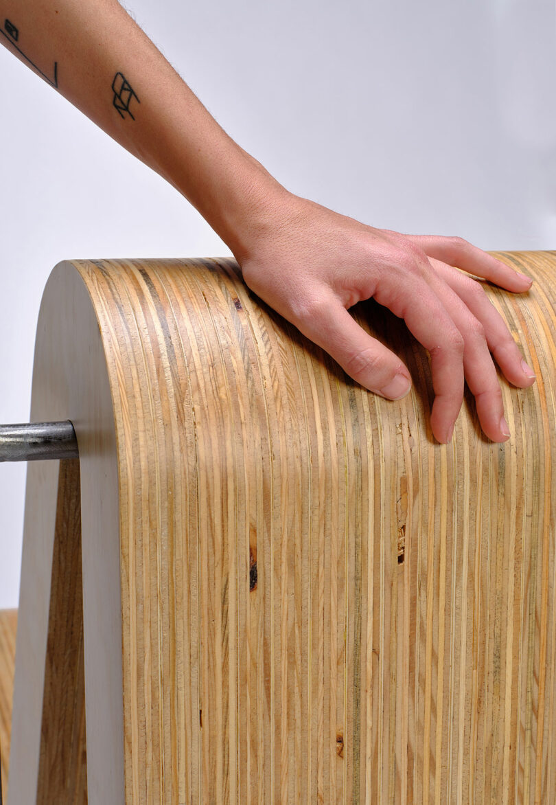 A hand rests on the curved edge of a layered plywood structure with a metal rod passing through it.