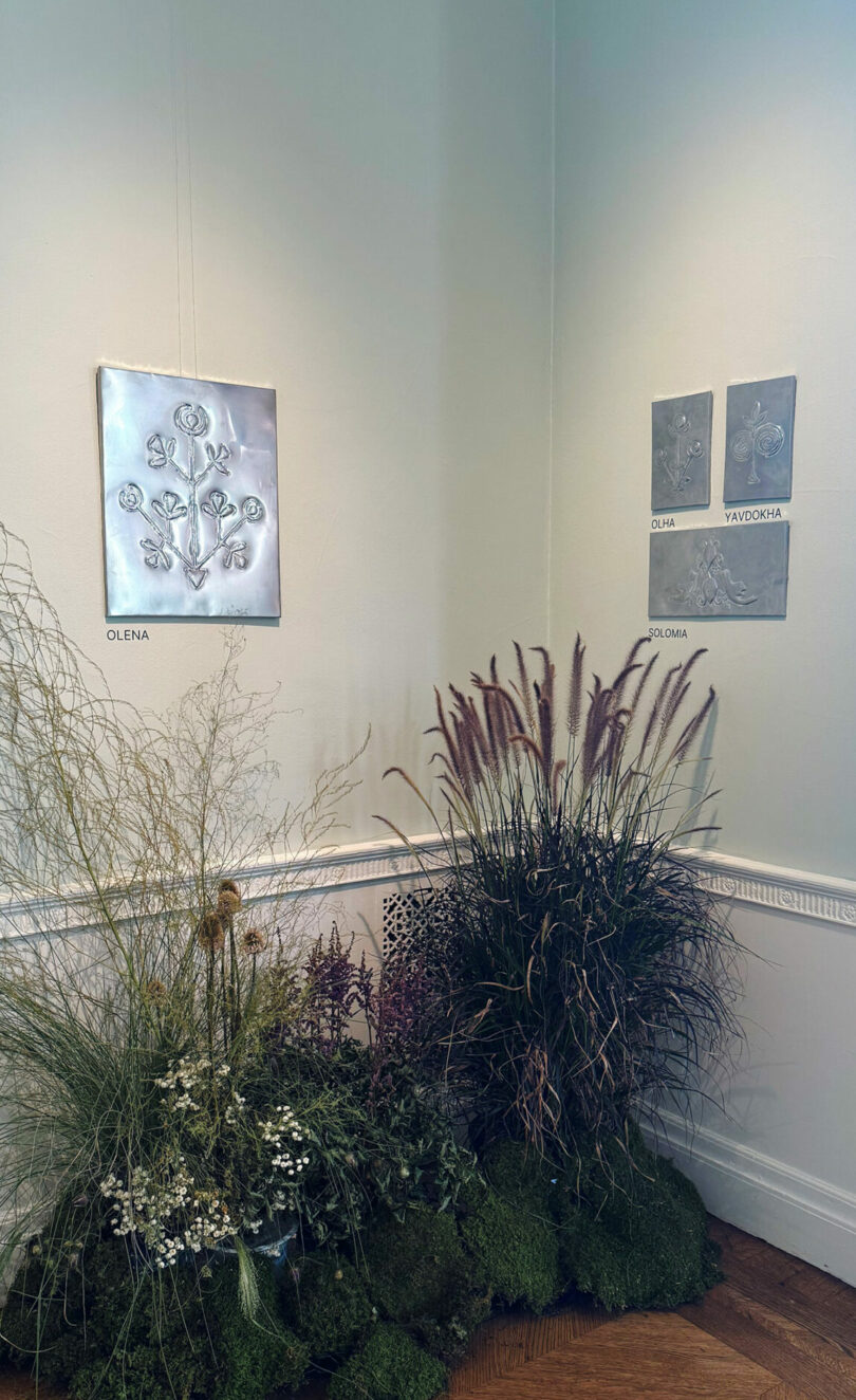 Art exhibit corner with metallic botanical reliefs labeled "OLENA," "YULIA," "YAVDOSIA," and "OKSANA" on pale walls above a Take 5-inspired display of ornamental grasses and greenery.