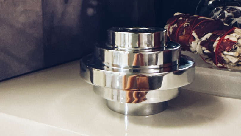 A shiny, stacked metal object with a cylindrical shape sits on a beige surface next to a bundle of dried herbs wrapped in red string, offering a moment to Take 5 and enjoy the calming ambiance.