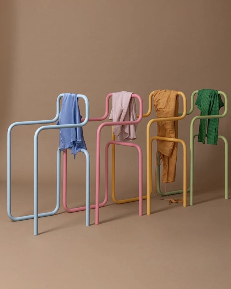 Four colorful, abstract, metal clothing racks stand in a row (blue, pink, yellow, and green), each holding a different item of clothing against a plain brown background