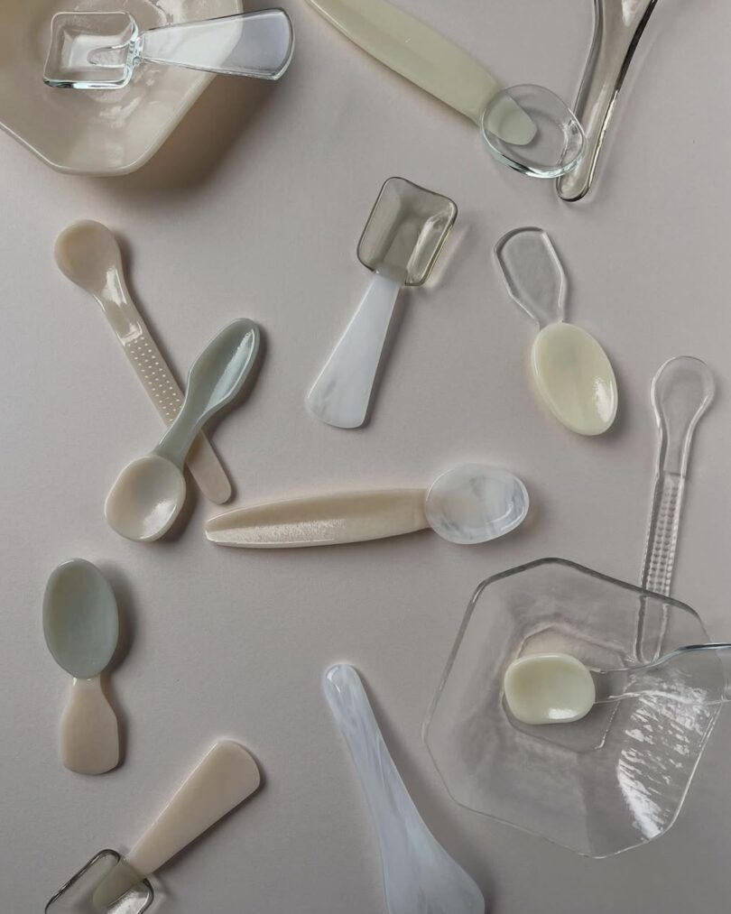 Assorted translucent and opaque glass spoons and dishes are arranged on a light beige surface.